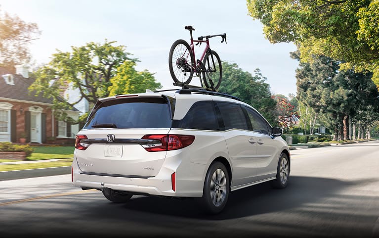 2027 Honda Odyssey Elite in Platinum White Pearl back driving through a sunny residential neighborhood (Minivan).